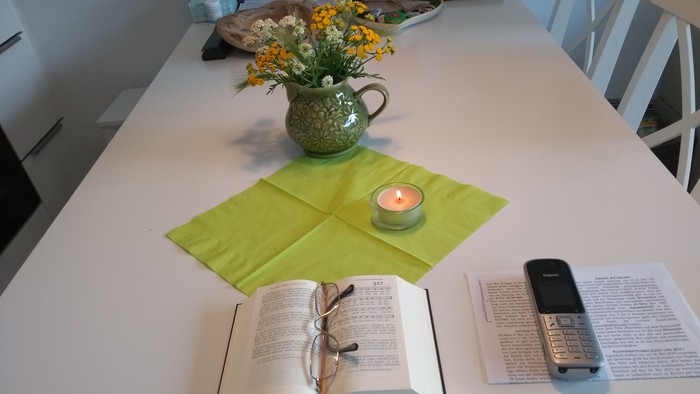 Eine ruhige Tischdekoration mit einem offenen Buch, einer Kerze, Blumen in einem Krug und einem alten Handy neben Notizen.