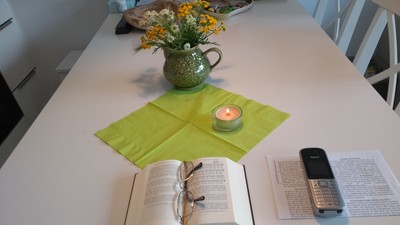 Eine ruhige Tischdekoration mit einem offenen Buch, einer Kerze, Blumen in einem Krug und einem alten Handy neben Notizen.