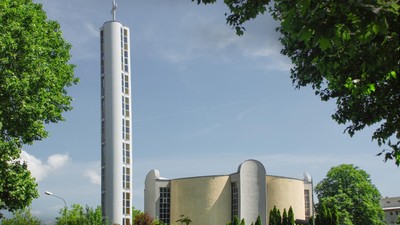 Moderne Kirche mit hohem Glockenturm, eingerahmt von üppigen grünen Bäumen unter einem teilweise bewölkten Himmel.