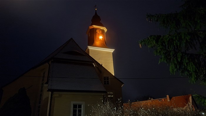 Ein beleuchteter Glockenturm einer Kirche bei Nacht, umgeben von Bäumen und einem dunklen Himmel.