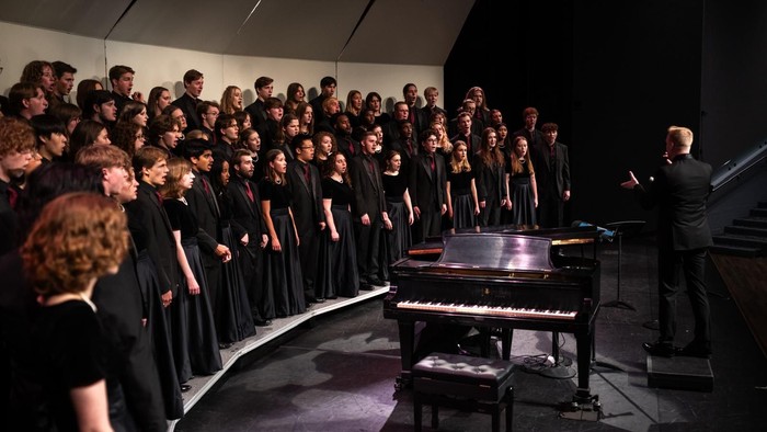 Ein großer Chor tritt auf der Bühne auf, begleitet von einem Dirigenten und einem Pianisten.