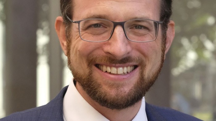 Ein Mann mit Brille und Bart, der einen Anzug und Krawatte trägt, lächelt in die Kamera.