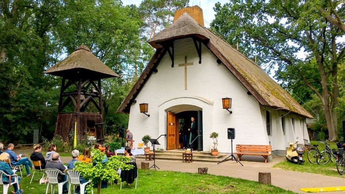 Kleine weiße Kirche mit Strohdach, umgeben von Bäumen und Menschen, die sich im Freien aufhalten.
