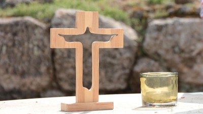 Ein hölzernes Kreuz und ein Glas mit Flüssigkeit stehen auf einer weißen Oberfläche vor einem felsigen Hintergrund