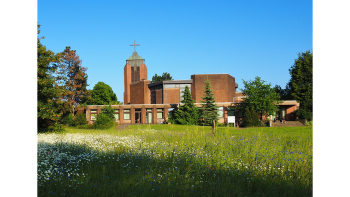 Ein rotes Backsteingebäude mit Turm und Kreuz, umgeben von grünem Gras und Bäumen unter einem klaren blauen Himmel
