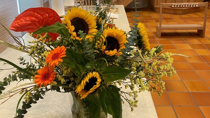 Lys blomsterdekoration i en vase på et kirkealter med lys og træbænke i baggrunden.