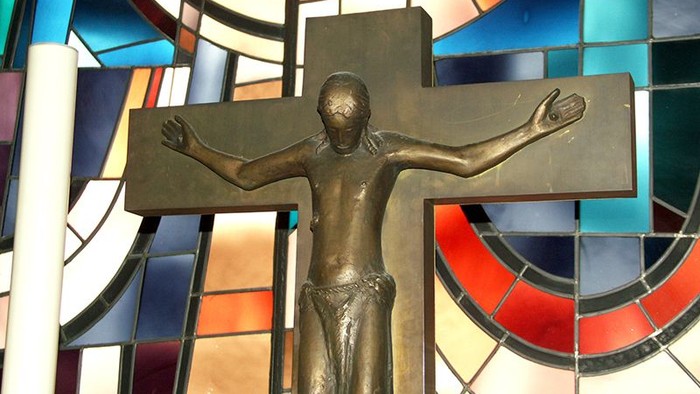 Bronzene Kruzifix-Skulptur mit ausgebreiteten Armen vor farbigem Kirchenfenster-Hintergrund.