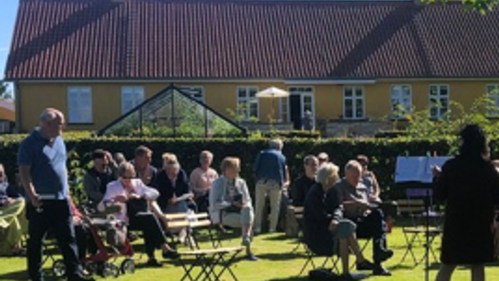 En gruppe af mennesker sidder ude og spiser på en grønne eng med et hus i baggrunden
