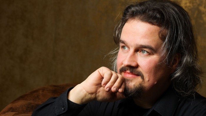 Ein Mann mit langem Haar und Bart sitzt nachdenklich auf einem braunen Sofa.