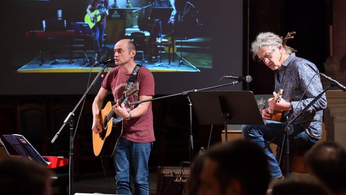 Deux hommes jouent de la guitare sur scène, accompagnés d'un public.