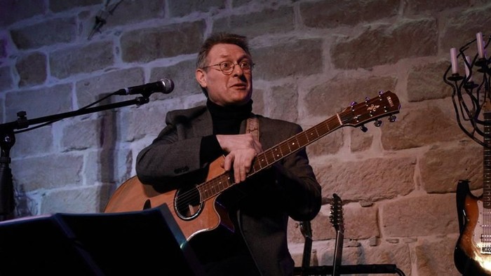 Ein Mann spielt akustische Gitarre auf der Bühne in einem rustikalen Veranstaltungsort mit Steinwänden.
