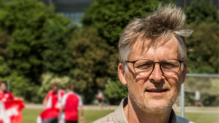 En mand med briller og kort hår står i en park foran et stort stadion.