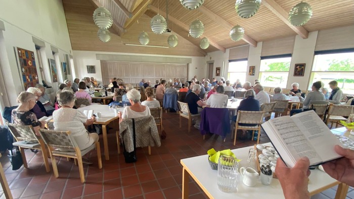Gruppen samles indendørs til et fælles måltid eller sammenkomst med åbne bøger og varmt lys.