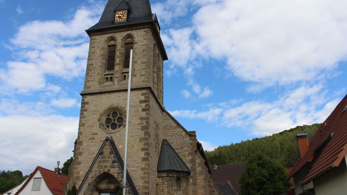 Kirche mit hohem Steinturm und Uhr, umgeben von Häusern und Bäumen unter blauem Himmel.