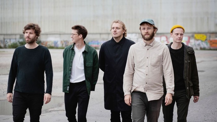 Fem mænd i afslappet påklædning, der går sammen i et urbant industriområde med graffitimalede vægge.