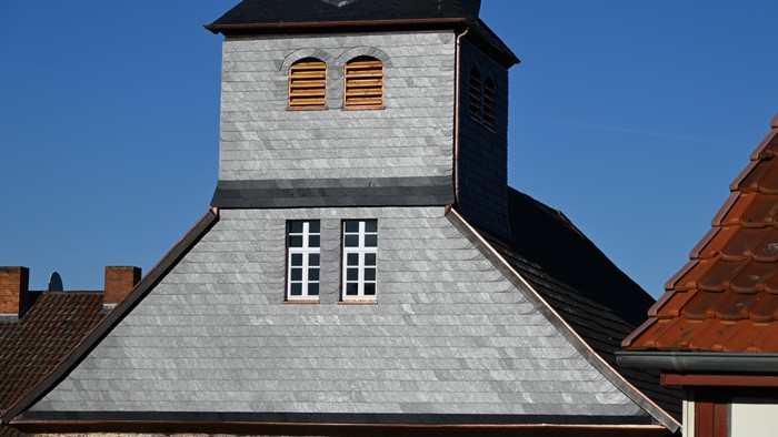 Graues Steingebäude mit kleinen Fenstern und einem steilen Dach unter einem klaren blauen Himmel.
