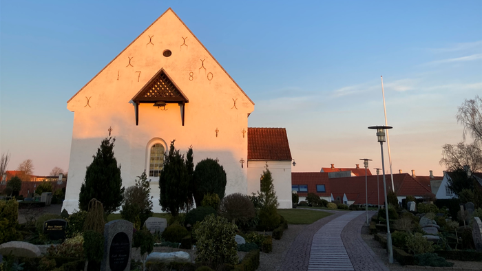 En lille, belyset kirke under en blå himmel med en halvmåne og fugle.