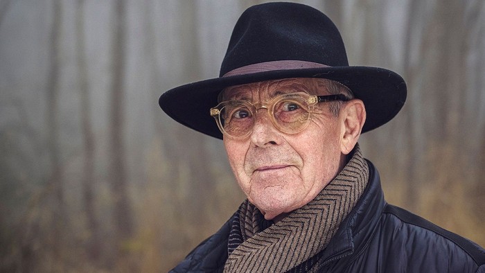 En ældre mand med fedora og briller står i en skovmiljø.