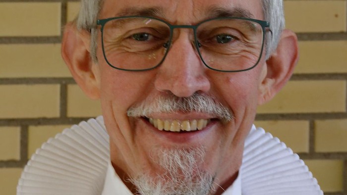 En smilende, eldre mand med hvid krage og briller.