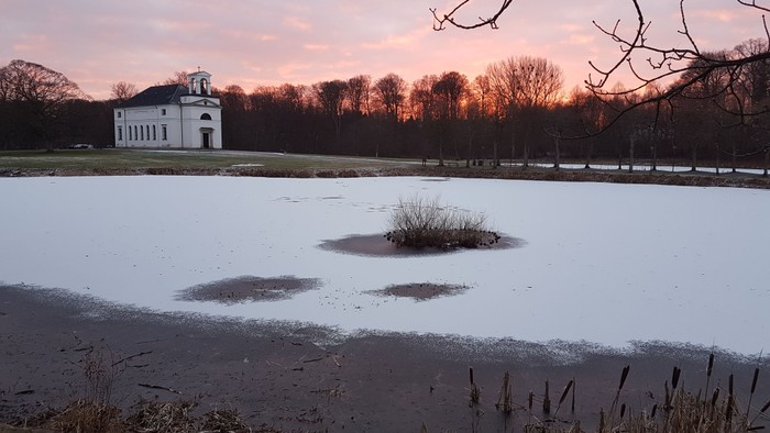 En fryset sø med en lille ø i midten og et hvidt hus i baggrunden ved solnedgang