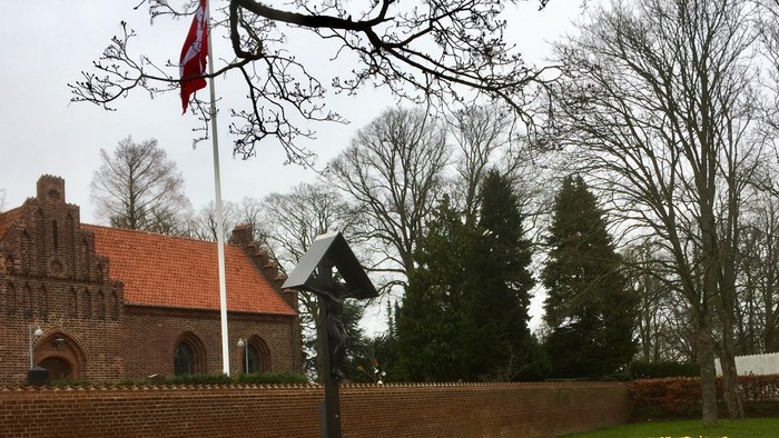 Historisk murstensbygning med flagstang og mindesmærke i et anlagt haveområde.