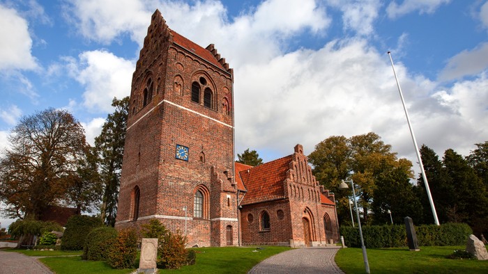 En rød murstenskirke med tårn og spiret, omgivet af græsplæne og træer under en blå Himmel med hvidt skyer