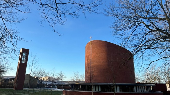 En moderne bygning med en rund rød fasade og flade tag, omgivet af træer og en klar blå himmel.