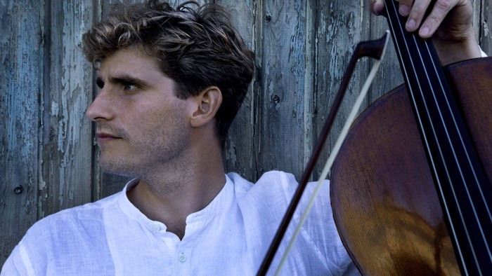 En mand i hvid skjorte, der spiller cello udendørs foran en træbaggrund.