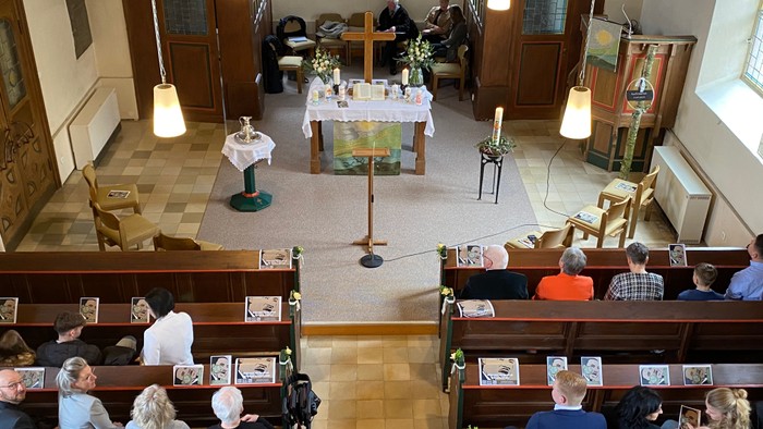 Kircheninneres mit Menschen, die in Kirchenbänken sitzen, Altar vorne mit Kreuz und Kerzen.