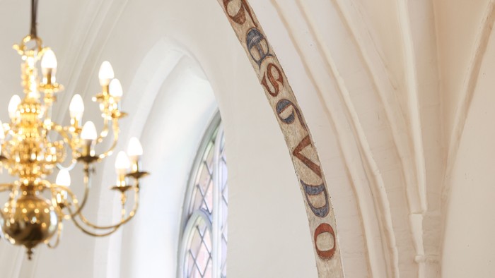 En belysning hænger fra loftet i en kirke med et bogstaveligt maleri på væggen.