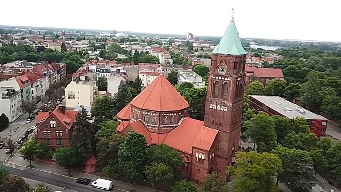 Luftaufnahme einer historischen Backsteinkirche mit hohem grünem Turm in einer grünen, städtischen Umgebung.