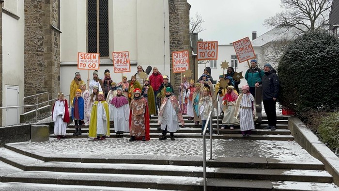 Eine Gruppe in bunten Kostümen marschiert vor einer Kirche und hält Protestschilder hoch, die sichere Versammlungen fordern.