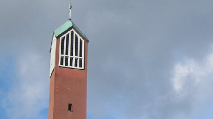 Eine rote Backsteinkirche mit einem hohen Glockenturm und Solarpaneelen auf dem Dach, umgeben von Grünflächen.
