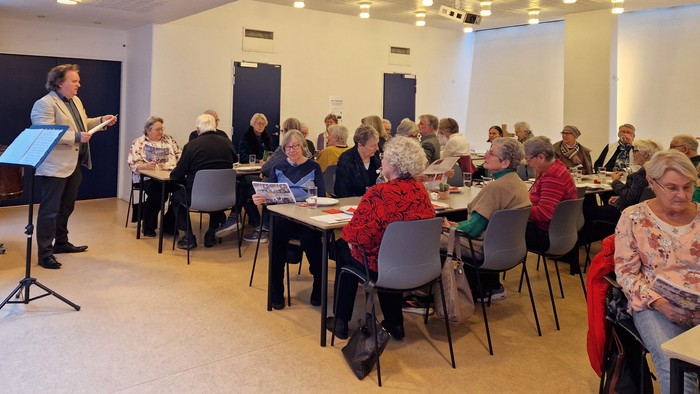 En kvinde står og taler til en gruppe af mennesker, der sidder ved bord i et lys og rummeligt lokal.