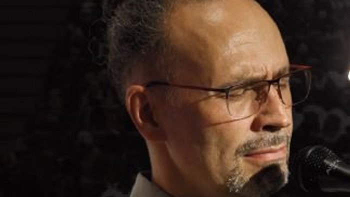 Ein Mann mit Brille und Bart, der auf einer Bühne in ein Mikrofon spricht.