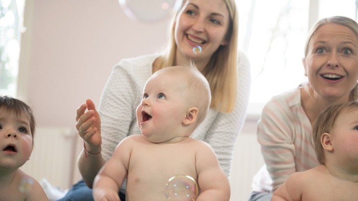 Ein Baby sitzt auf dem Schoß einer Frau, umgeben von anderen Babys und einer Frau, die lacht.
