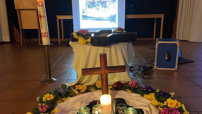 Auf dem Boden des Saals steht ein Holzkreuz in der Mitte eines Kreises mit brennenden Kerzen und Blumen.