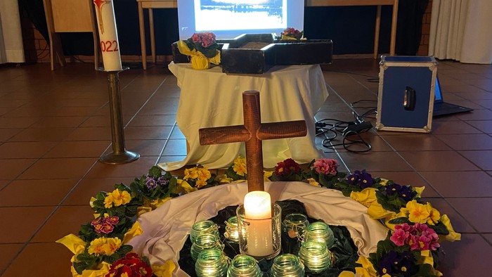 Auf dem Boden des Saals steht ein Holzkreuz in der Mitte eines Kreises mit brennenden Kerzen und Blumen.