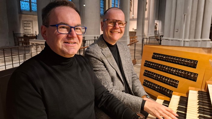 Zwei Männer lächeln neben einer großen Kirchenorgel in einem prunkvollen Innenraum.