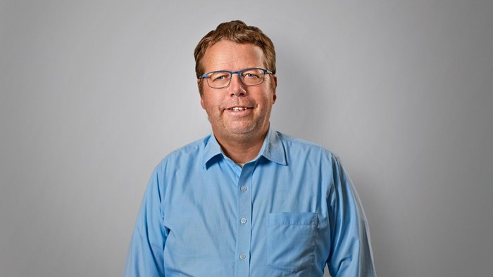 En mand med briller og blå skjorte står foran en grå væg.