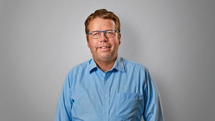 En mand med briller og blå skjorte står foran en grå væg.