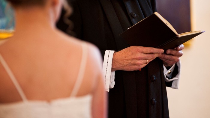 En person i bryllupsantræk ser til, mens en vielsesleder læser op fra en bog under en ceremoni.