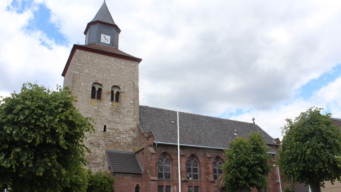 Steinerne Kirche mit hohem Uhrturm und Rundbogenfenstern, umgeben von Bäumen.