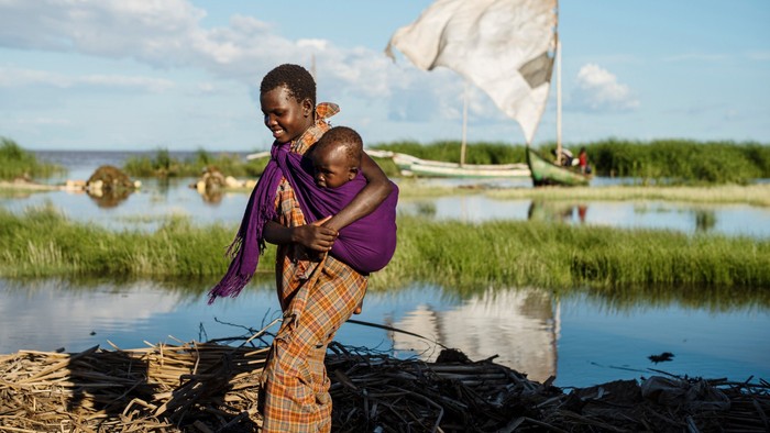 En kvinde med barn på armene står ved vandet med en sejlbåd i baggrunden