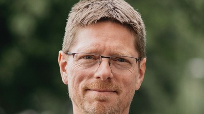 En mand med briller og stribet trøje står i en grøn og udefineret naturmiljø.