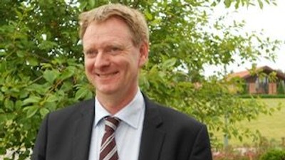 En smilende mand i jakkesæt og slips, der poser udendørs mellem grønt løv.