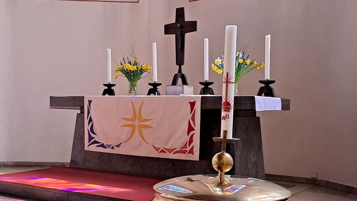 Eleganter Kirchenaltar mit Kerzen, Blumen und einem goldenen Taufbecken vor einem verzierten Tuch.