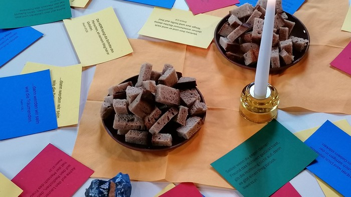 Bunte Zettel mit Texten, zwei Schalen mit Brot und eine brennende Kerze auf Papiertischdecke