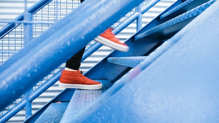 Person steigt eine blaue Metalltreppe hinauf, trägt rote Turnschuhe
