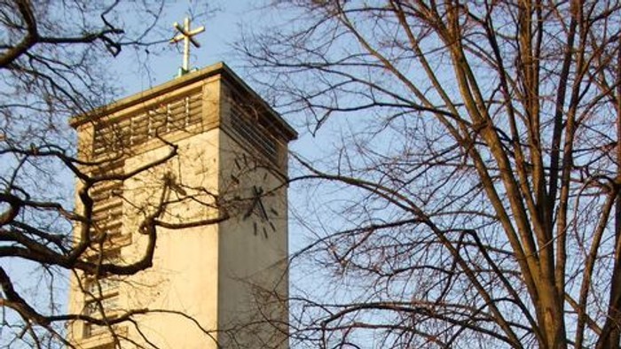 Ein hoher Turm mit Uhr und Kreuz vor kahlen Bäumen